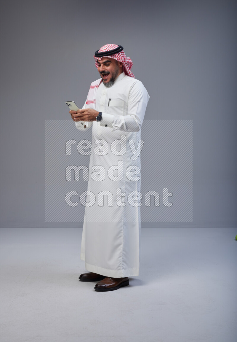 Saudi man Wearing Thob and red Shomag standing texting on phone on Gray background