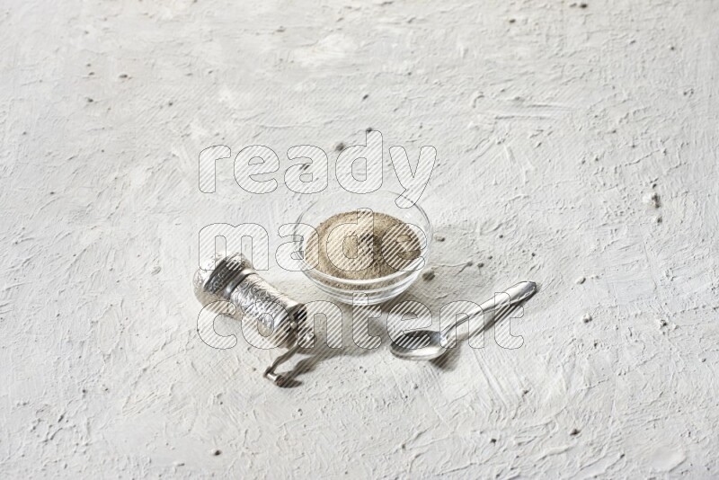 A glass bowl full of white pepper powder with white pepper beads and a metal grinder on textured white flooring