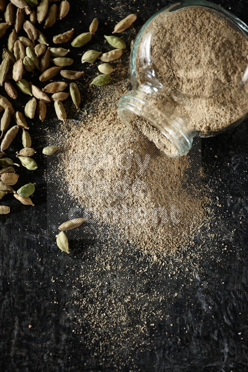 وعاء زجاجي للتوابل مقلوب وممتلئ ببودرة الهيل والبودرة تناثرت منه مع حبوب الهيل المتناثرة على خلفية سوداء