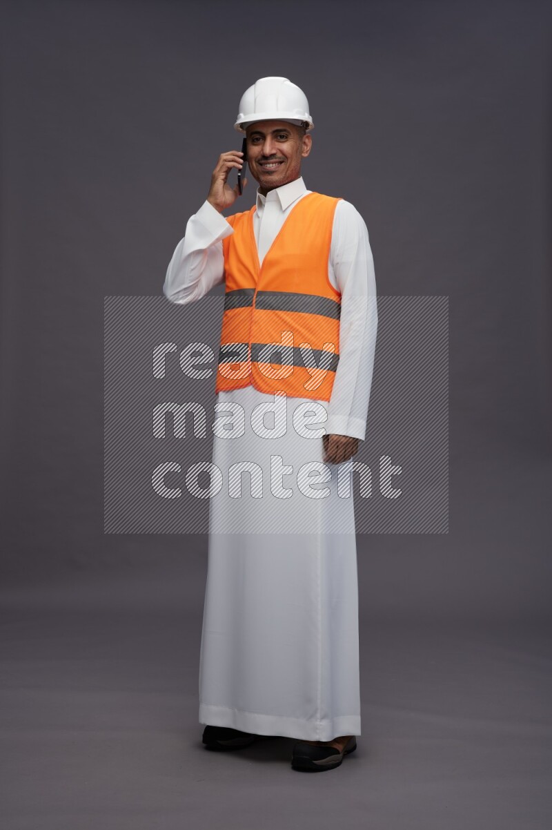 Saudi man wearing thob with engineer vest standing talking on phone on gray background