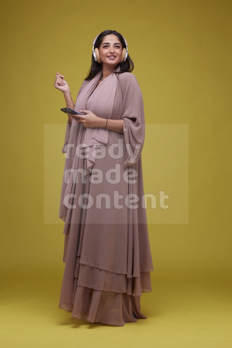 A Saudi woman Listing to Music on a Yellow Background wearing Brown Abaya