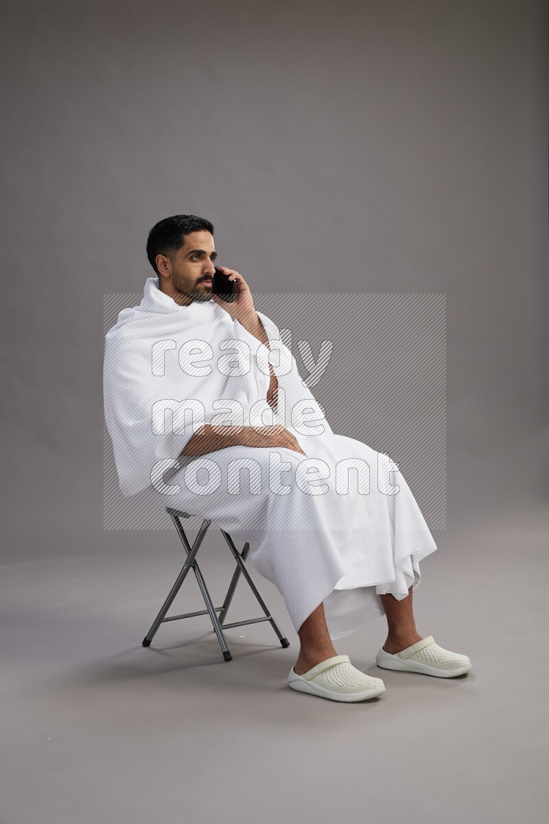 A man wearing Ehram sitting on chair talking on the phone on gray background
