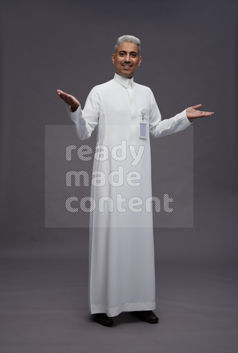 Saudi man wearing thob with pocket employee badge standing interacting with the camera on gray background