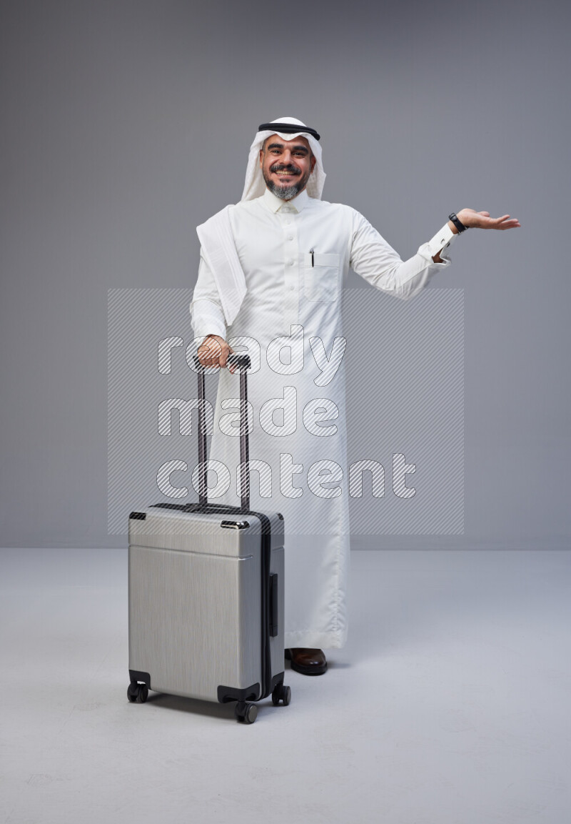 Saudi man wearing Thob and white Shomag standing holding Travel bag on Gray background