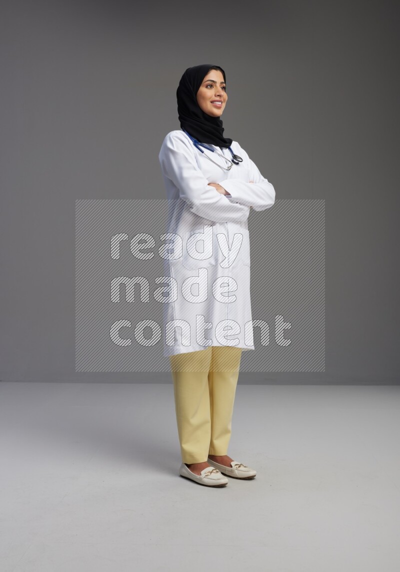 Saudi woman wearing lab coat with stethoscope standing with crossed arms on Gray background