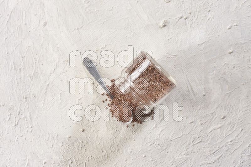 A glass spice jar full of flax seeds flipped with a metal spoon full of the seeds on a textured white flooring