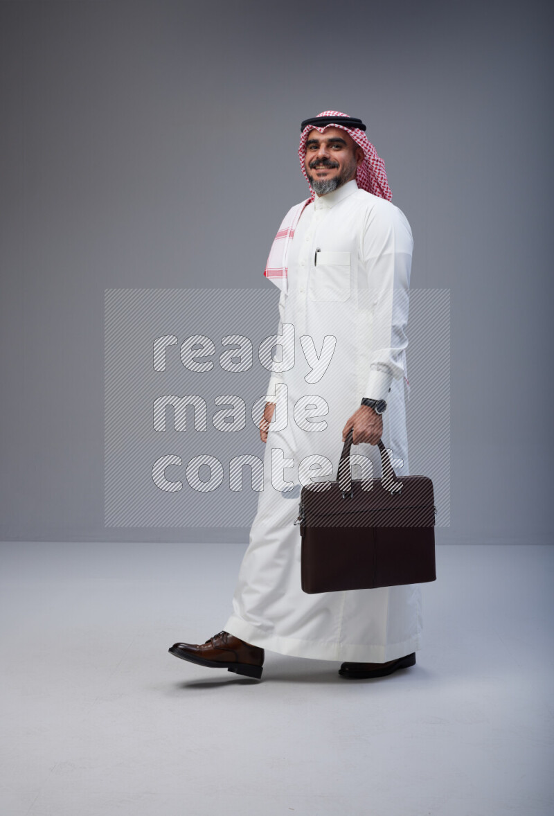 Saudi man Wearing Thob and red Shomag standing holding bag on Gray background