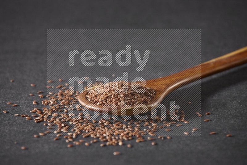 A wooden ladle full of flaxseeds and seeds spread beside it on a black flooring