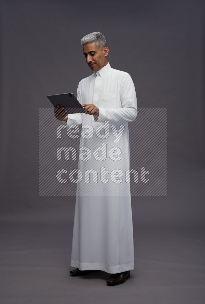 Saudi man wearing thob standing working on tablet on gray background