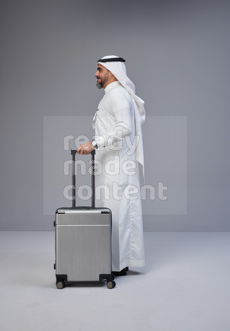 Saudi man wearing Thob and white Shomag standing holding Travel bag on Gray background