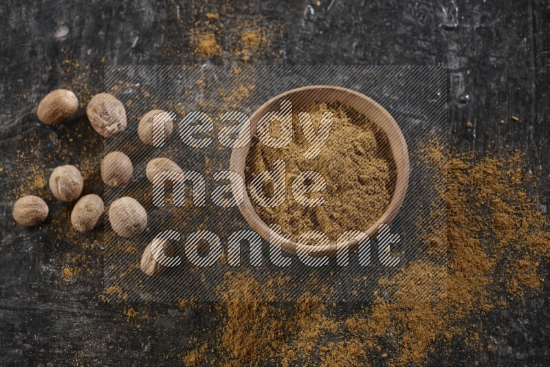A wooden bowl full of nutmeg powder with whole seeds and sprinkled powder beside it on a textured black flooring