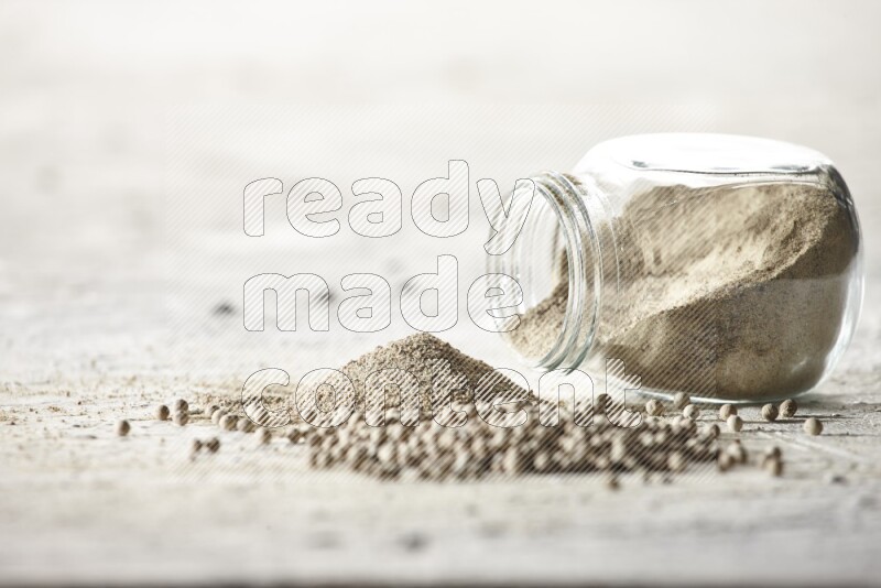 A flipped herbal glass jar full of white pepper powder with spilled powder and beads on textured white flooring
