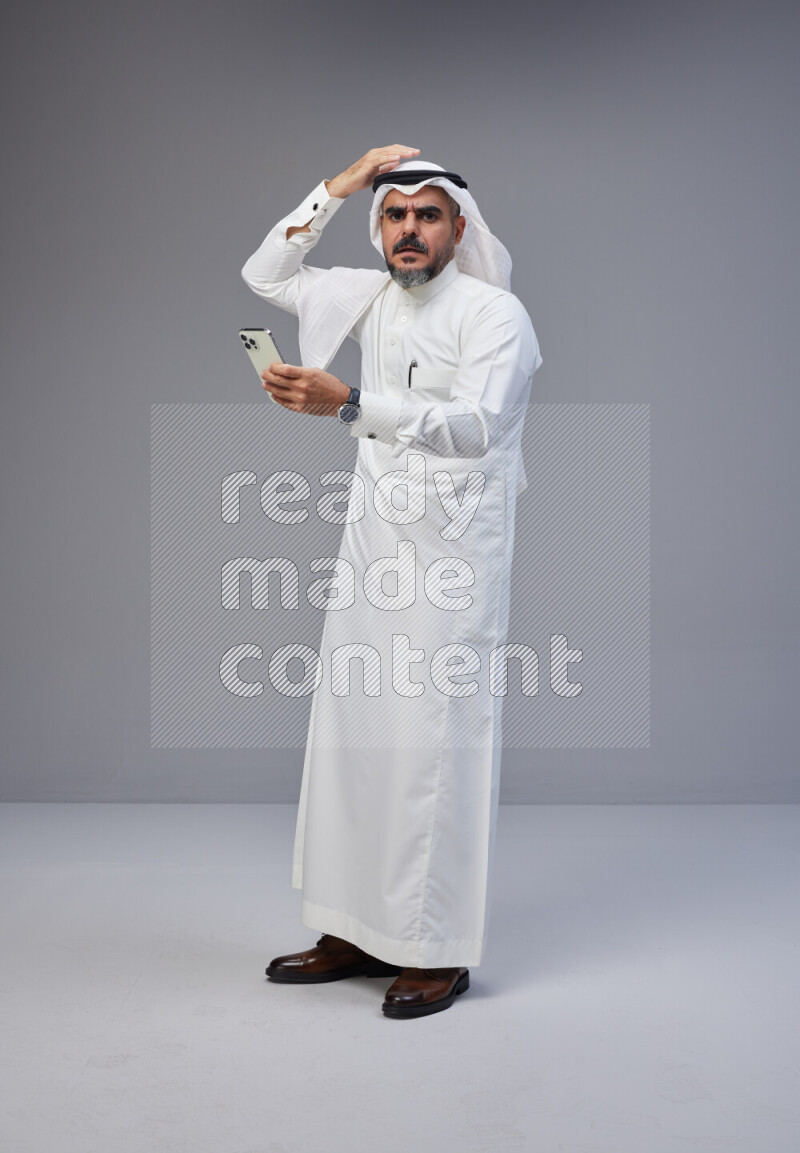 Saudi man Wearing Thob and white Shomag standing texting on phone on Gray background