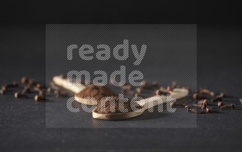 ملعقتان خشبيتان ممتلئتان ببودرة القرنفل مع حبوب القرنفل المتناثرة على خلفية سوداء