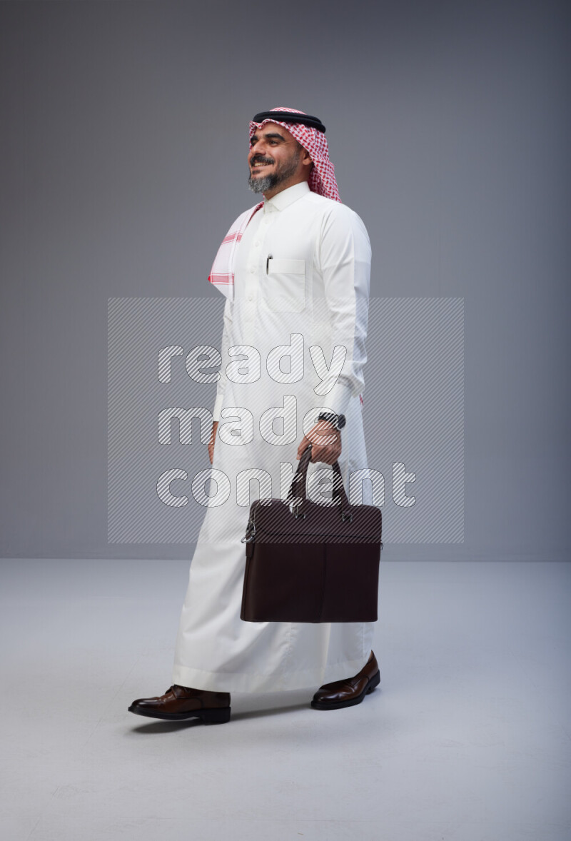Saudi man Wearing Thob and red Shomag standing holding bag on Gray background