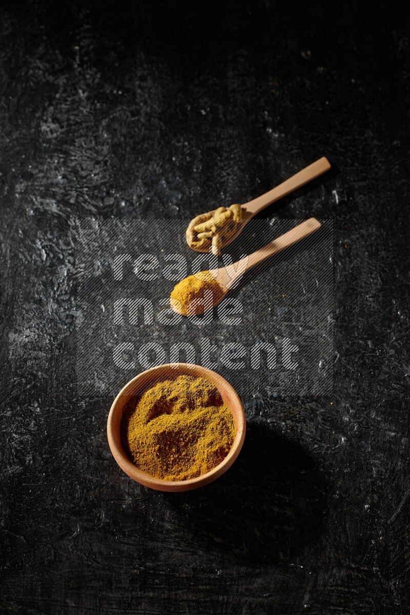 A wooden bowl full of turmeric powder and 2 wooden spoons full of dried turmeric finger and turmeric powder on textured black flooring