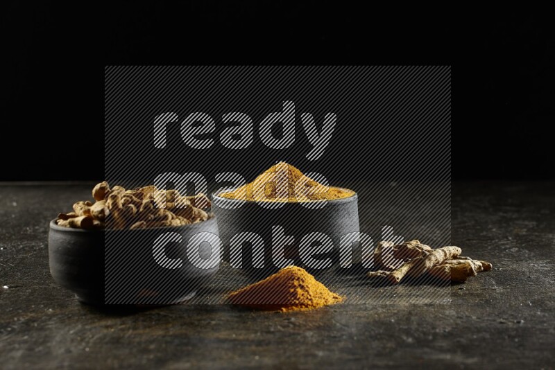 2 pottery black bowls, one full of turmeric powder and the other full of dried turmeric whole fingers on a textured black flooring