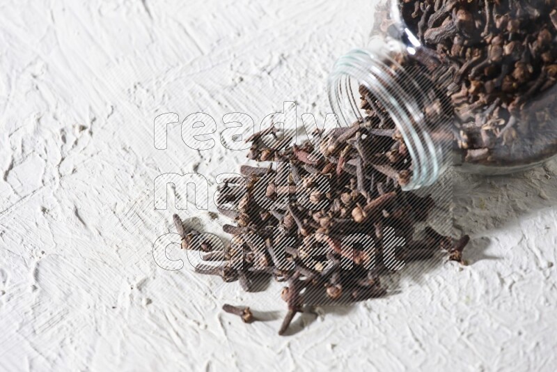 A flipped glass spice jar full of cloves and cloves came out of it on textured white flooring