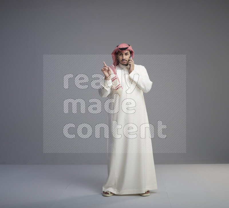 A Saudi man standing wearing thob and red shomag talking on phone on gray background