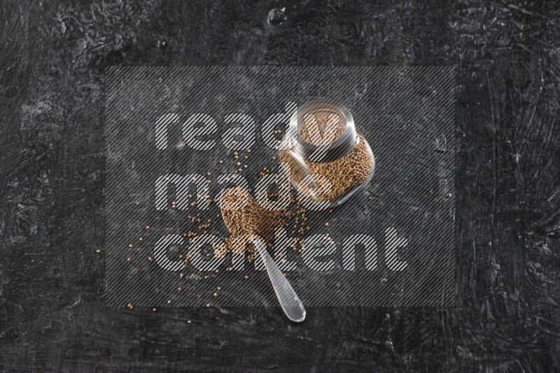 A glass spice jar and a metal spoon full of mustard seeds on a textured black flooring