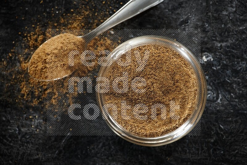 A glass jar and a metal spoon full of cumin powder on a textured black flooring