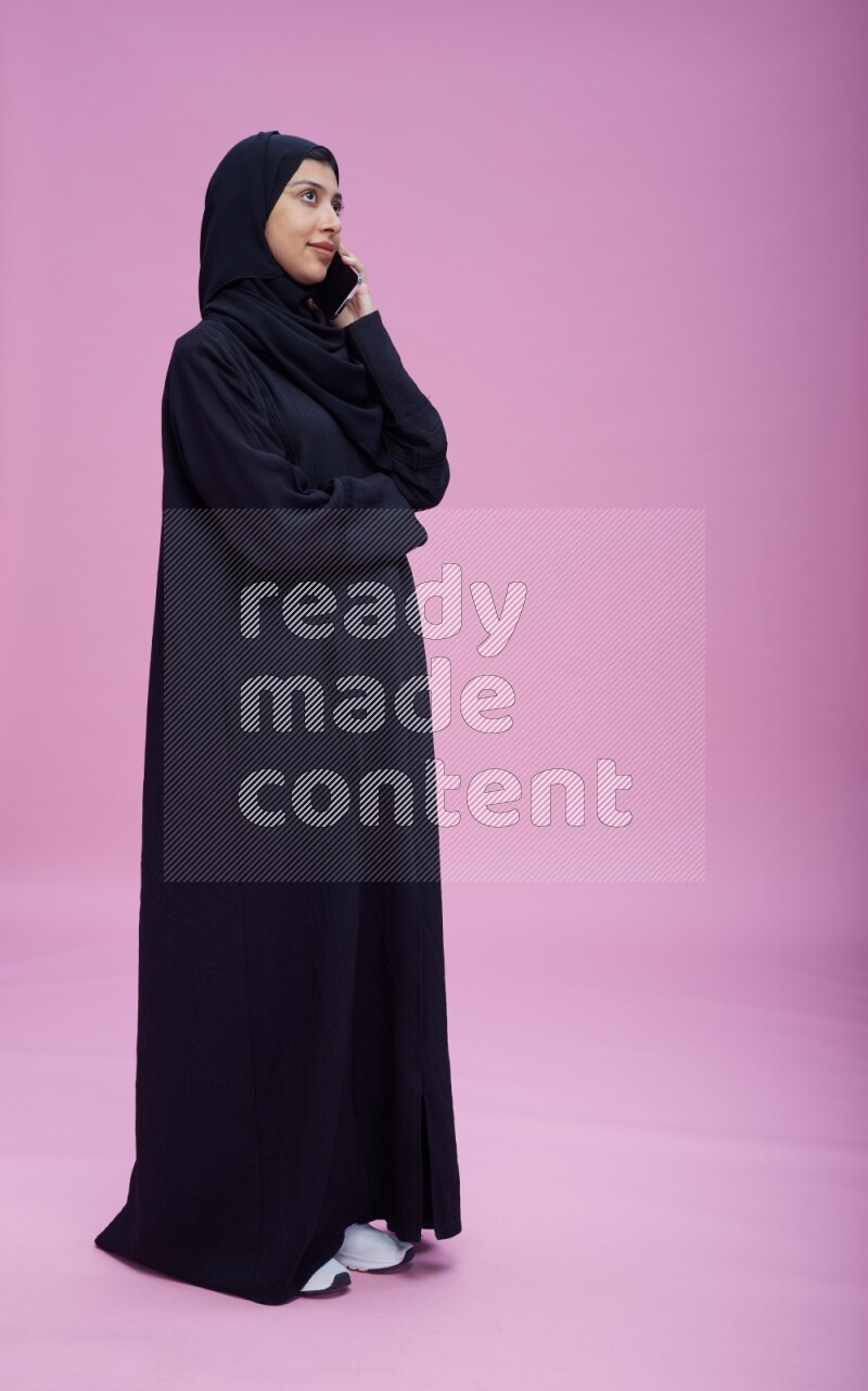 Saudi woman wearing Abaya standing talking on phone on pink background