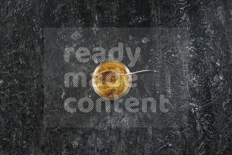 A glass jar and a metal spoon full of turmeric powder on a textured black flooring