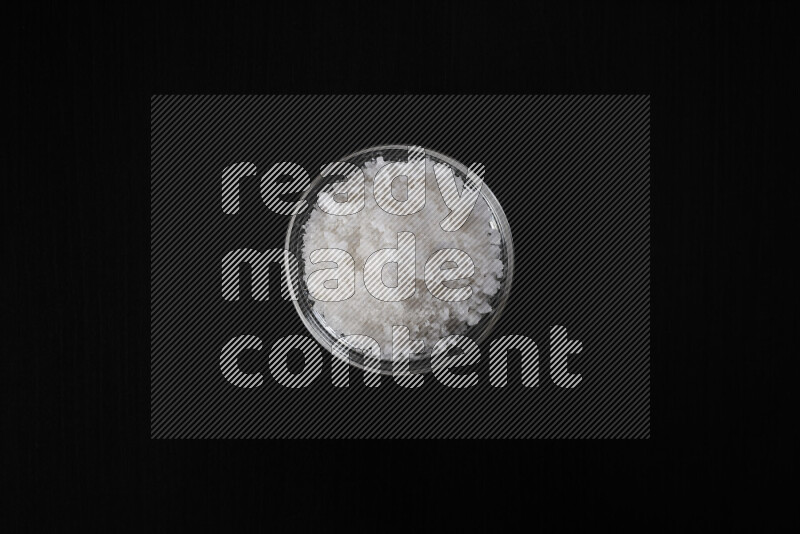 A glass bowl full of coarse sea salt crystals on black background