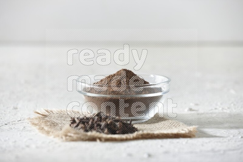 A glass bowl full of cloves powder and cloves grains on a burlap piece on a white flooring