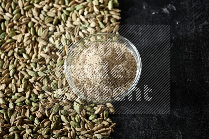 A glass bowl full of cardamom powder and cardamom seeds spreaded beneath the bowl on textured black flooring
