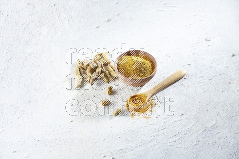 A wooden bowl and wooden spoon full of turmeric powder with dried turmeric fingers beside it on textured white flooring