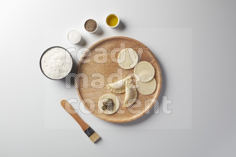 two closed sambosas and one open sambosa filled with meat while flour, salt, black pepper and oil with oil brush aside in a wooden dish on a white background
