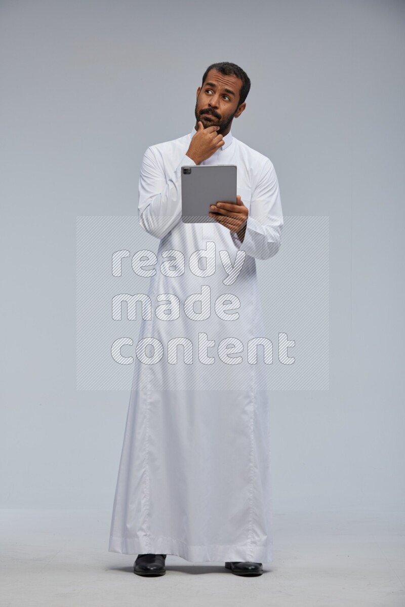Saudi man wearing Thob standing working on tablet on Gray background