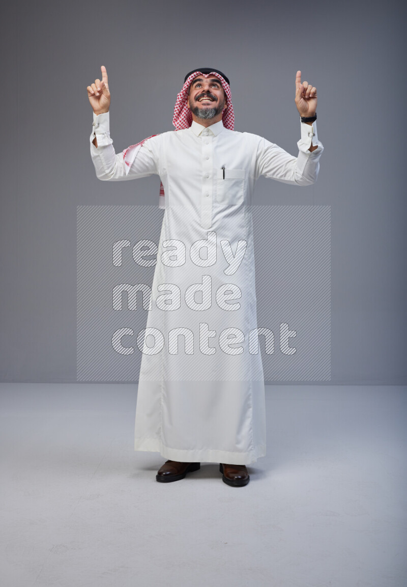 Saudi man Wearing Thob and red white Shomag standing interacting with the camera on Gray background
