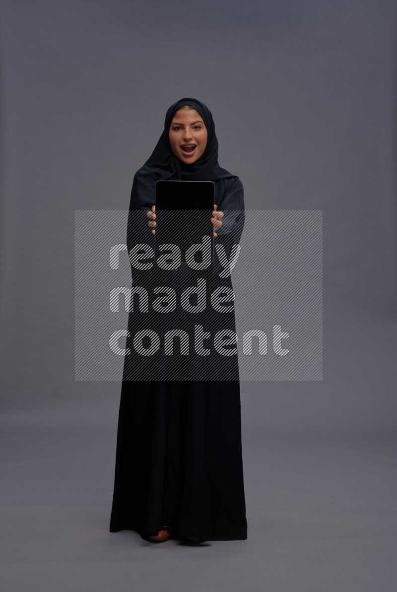 Saudi woman wearing Abaya standing showing tablet to camera on gray background
