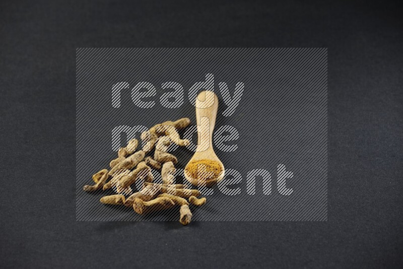 A wooden spoon full of turmeric powder with dried turmeric fingers on black flooring