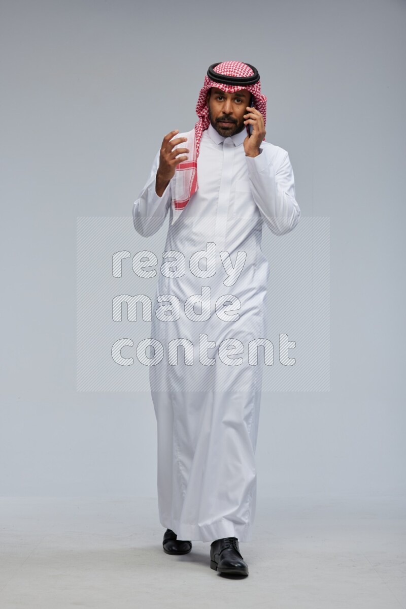 Saudi man Wearing Thob and shomag standing talking on phone on Gray background