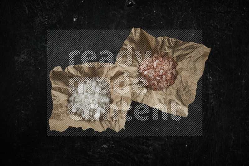 2 crumpled pieces of paper full of pink himalayan salt and white salt on black background