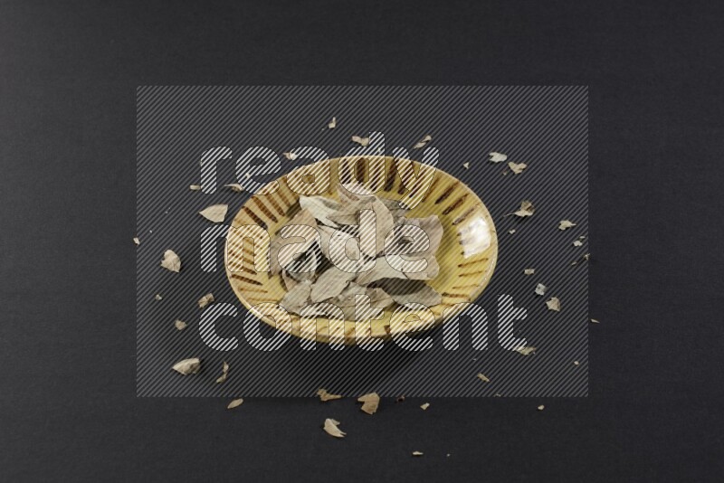 A multicolored pottery plate filled with dried bay leaves on black flooring