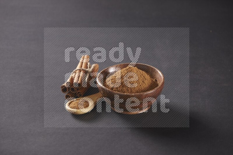 Cinnamon sticks stacked and bounded beside a wooden bowl full of cinnamon powder and a wooden spoon full of powder on black background