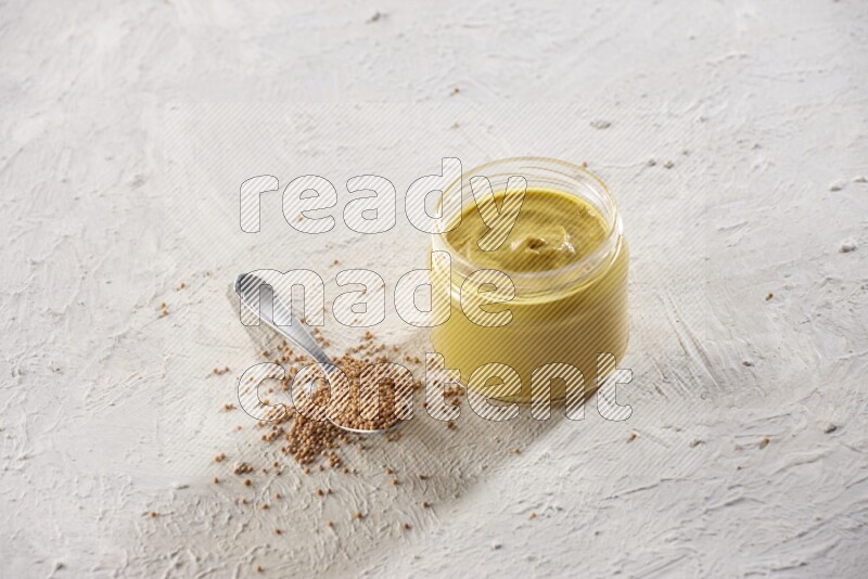 A glass jar full of mustard paste and a metal spoon full of mustard seeds on a textured white flooring