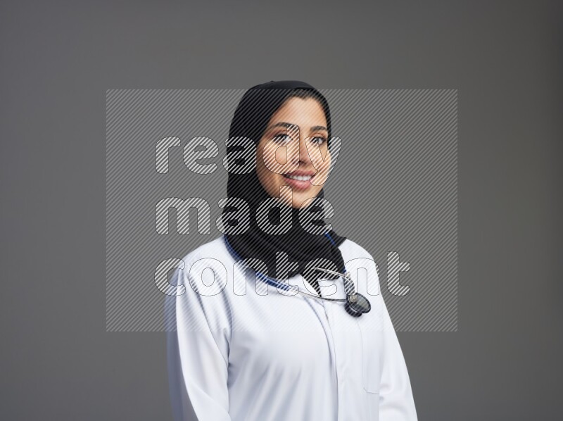 Saudi woman wearing lab coat with stethoscope standing interacting with the camera on Gray background