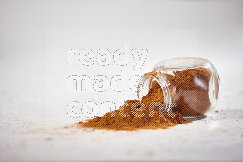 A glass jar full of ground paprika powder flipped with some spilling powder on white background