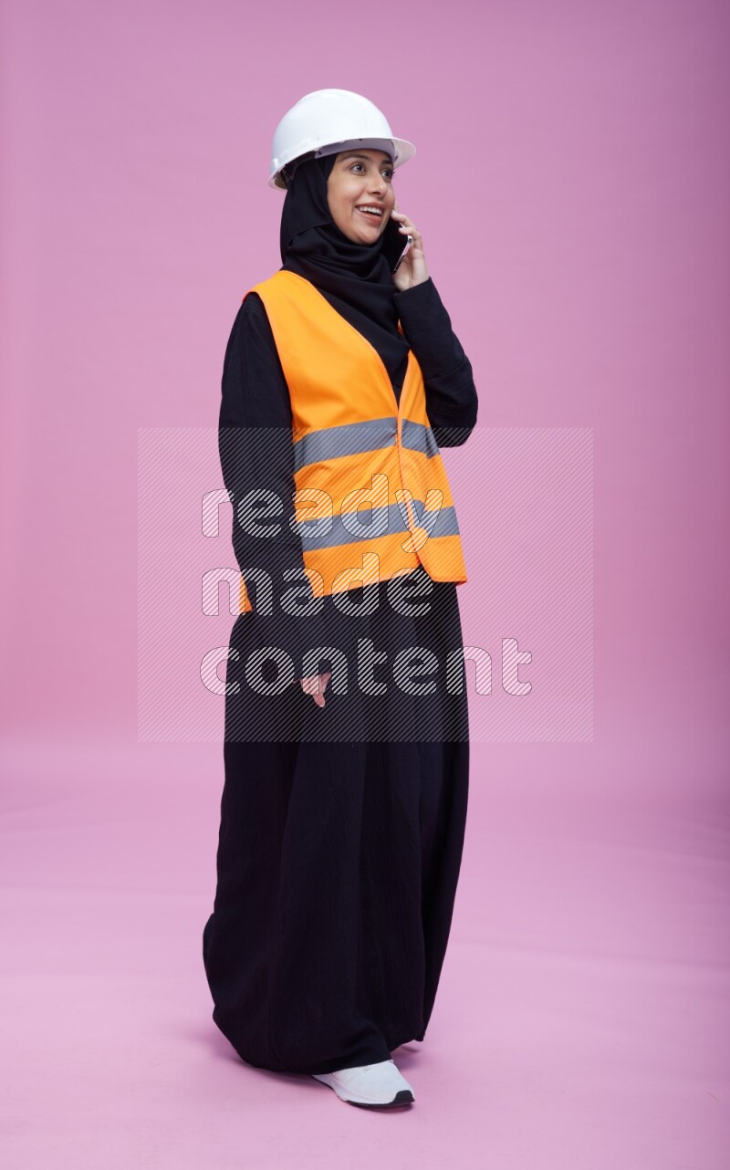 Saudi woman wearing Abaya with engineer vest and helmet standing talking on phone on pink background