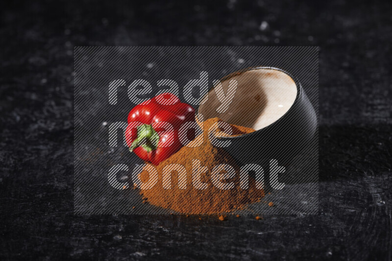 A black pottery bowl full of ground paprika powder with fallen powder from it on black background