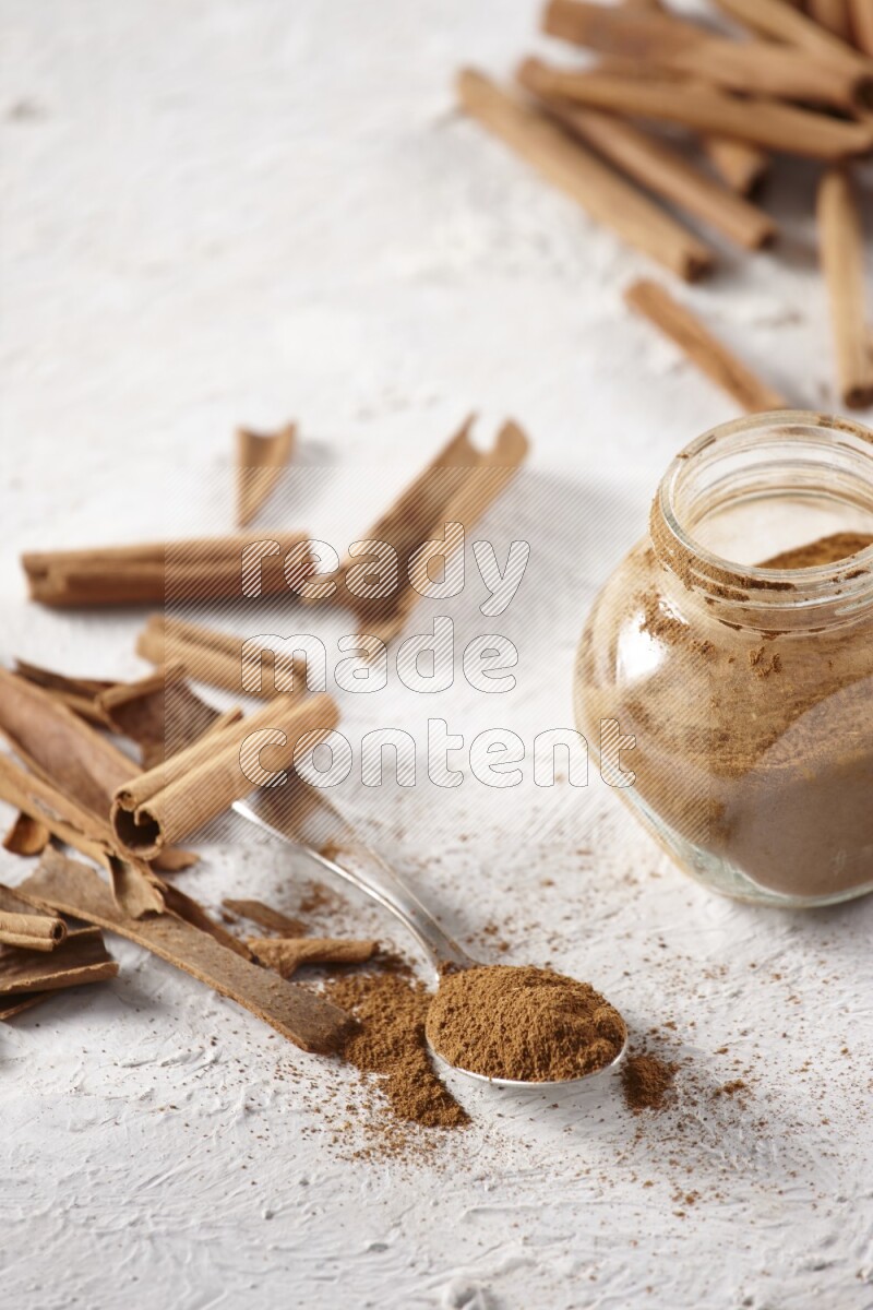 Herbal glass jar full cinnamon powder and a metal spoon surrounded by cinnamon sticks on a white background