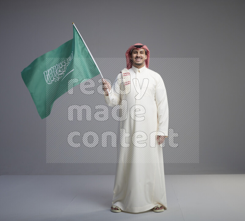 A Saudi man standing wearing thob and red shomag raising big Saudi flag on gray background