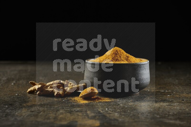 A pottery black bowl and a metal spoon full of turmeric powder with dried turmeric fingers on a textured black flooring