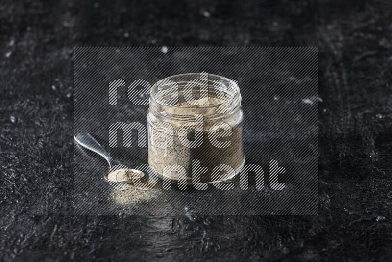 A glass jar and a metal spoon full of white pepper powder on textured black flooring