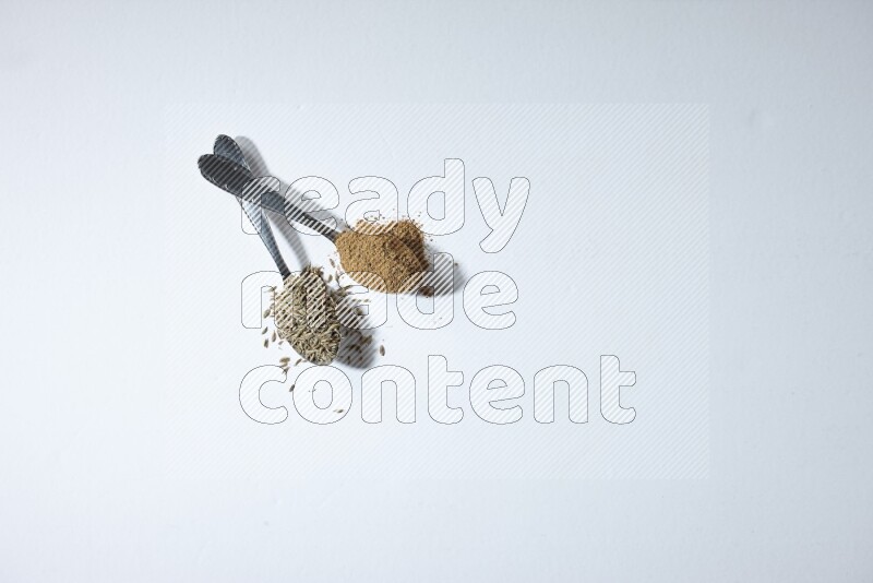 ملعقتان معدنيتان ممتلئتان بحبوب الكمون وبودرته على خلفية بيضاء